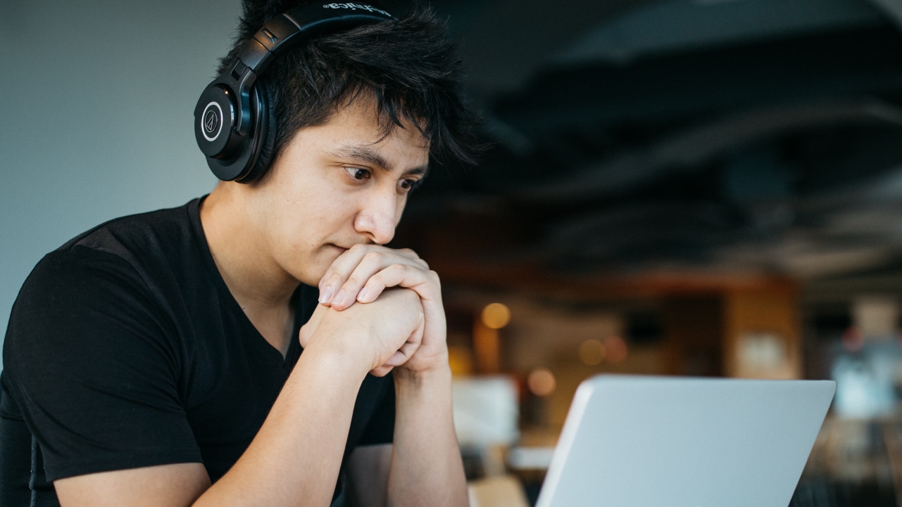 A young man works at a computer. He has headphones on, his hands are folded in front of him and held to his mouth.