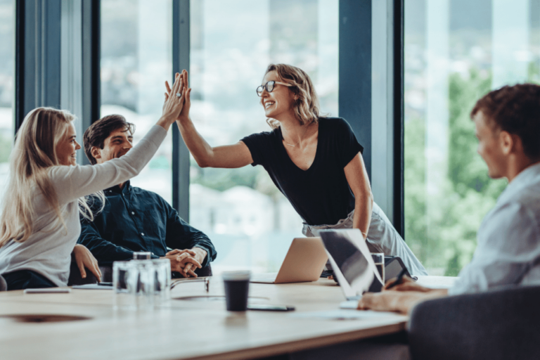 A female leader smiles and high fives a colleague across the table in a group meeting.